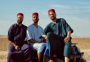 Three men wearing traditional fez hats pose confidently on a blue quad bike in a desert setting.