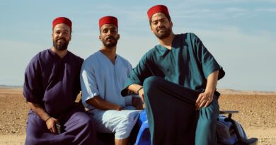 Three men wearing traditional fez hats pose confidently on a blue quad bike in a desert setting.