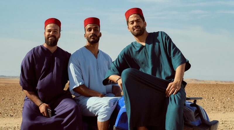 Three men wearing traditional fez hats pose confidently on a blue quad bike in a desert setting.